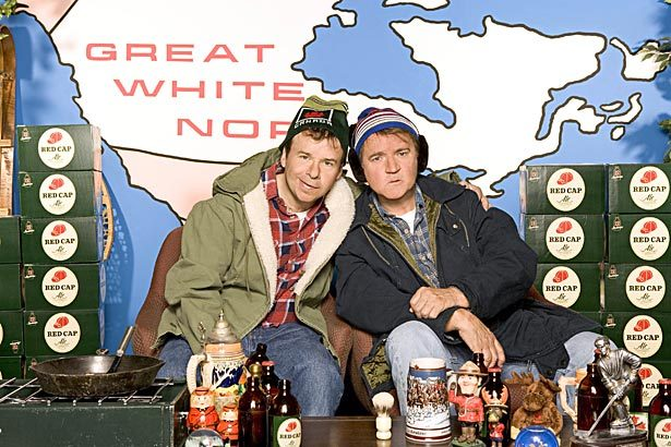 Photograph of the characters Bob and Doug Mackenzie. They are dressed in warm, rugged winter clothing—plaid shirts, parkas, and knit hats—sitting closely on a cozy set with Canadian-themed decor. Behind them is a map labeled “GREAT WHITE NORTH,” referencing Canada. The set is filled with cases of "Red Cap" beer, various knickknacks including beer steins, a moose figurine, a Mountie doll, and other rustic props.
