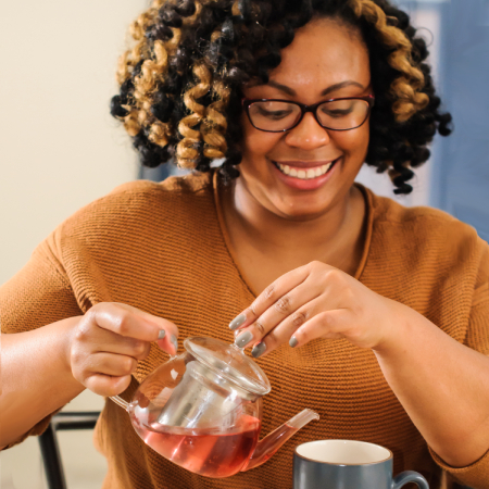 a Black femme person with gray nail polish wearing an orange sweater and smiling while pouring red tea into a mug from a glass teapot