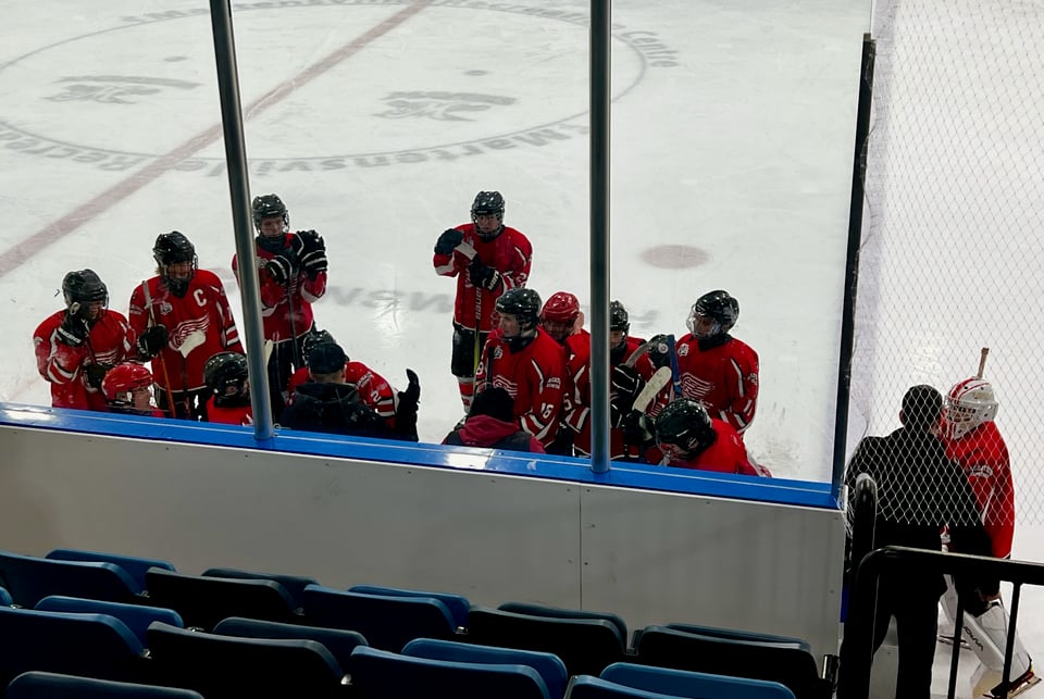 U18C hockey players gather around the bench on the ice between periods of a hockey game.