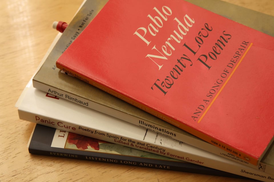 A small stack of poetry books rests on a wooden table, with Pablo Neruda’s Twenty Love Poems and a Song of Despair on top in its distinctive red cover. Beneath it are volumes including Arthur Rimbaud’s Illuminations, Panic Cure: Poetry from Spain for the 21st Century, and another partially visible collection. Soft lighting emphasizes the warm tones of the table and the worn, well-loved look of the books.