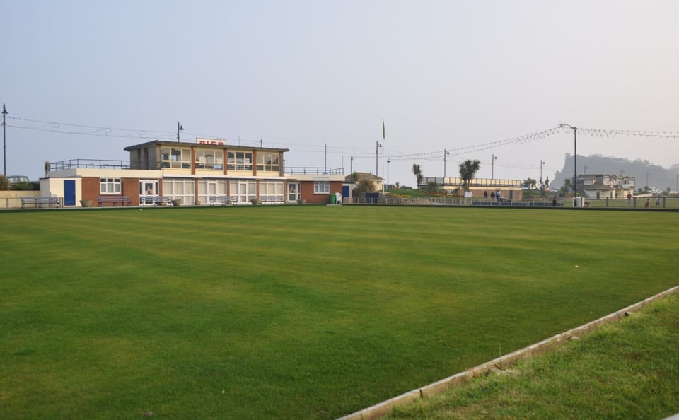 A large flat bowling green at a seafront. On the far side of it is a two-storey concrete and brick building. The lower level is has four sets of full length windows and doors, with single storey brick buildings on either side. On the upper level is a glass room with frames that all line up with the ground floor.