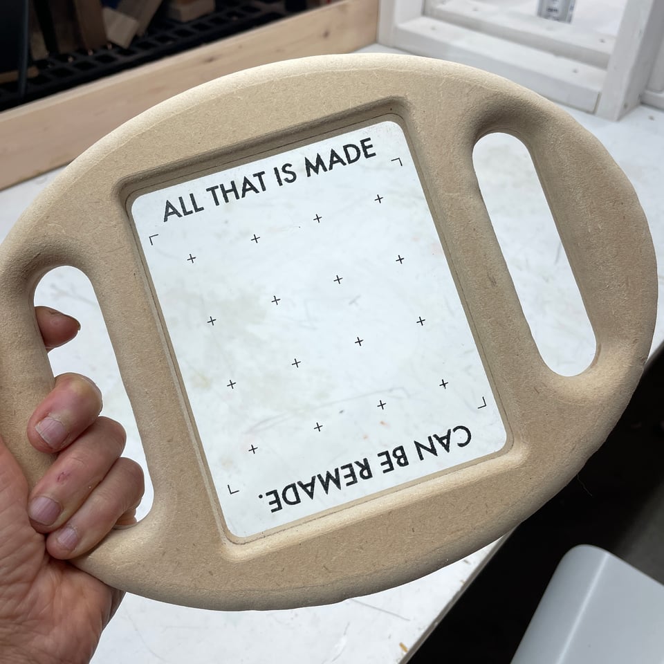 Hand holding an oval wooden frame, with cutout handles on each side (like a steering yoke). In the centre of the frame is a piece of clear glass, marked like a viewfinder, withe the words ALL THAT IS MADE across the top and CAN BE REMADE upside-down, across the bottom. The design suggests it can be held up to frame something in the environment.