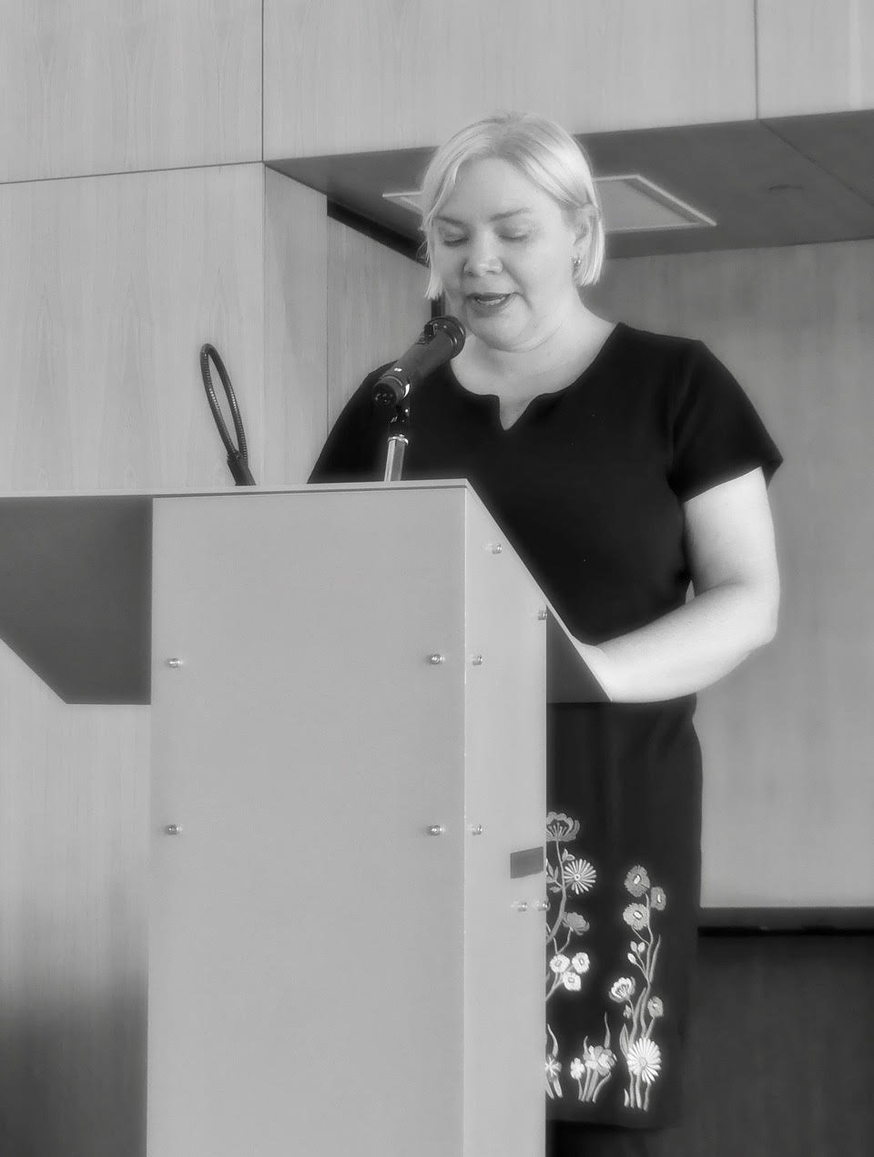 Black and white photo of Kate standing at a lectern and reading.