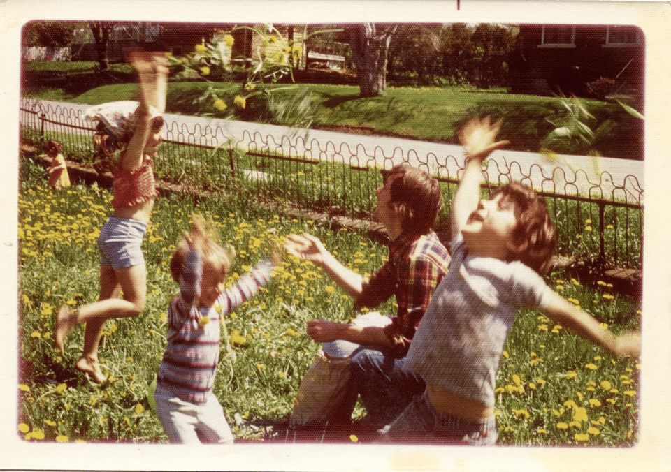 a vintage photo with young children playing in a grassy yard full of dandelions while an adult is kneeling down and tossing the dandelions in the air.