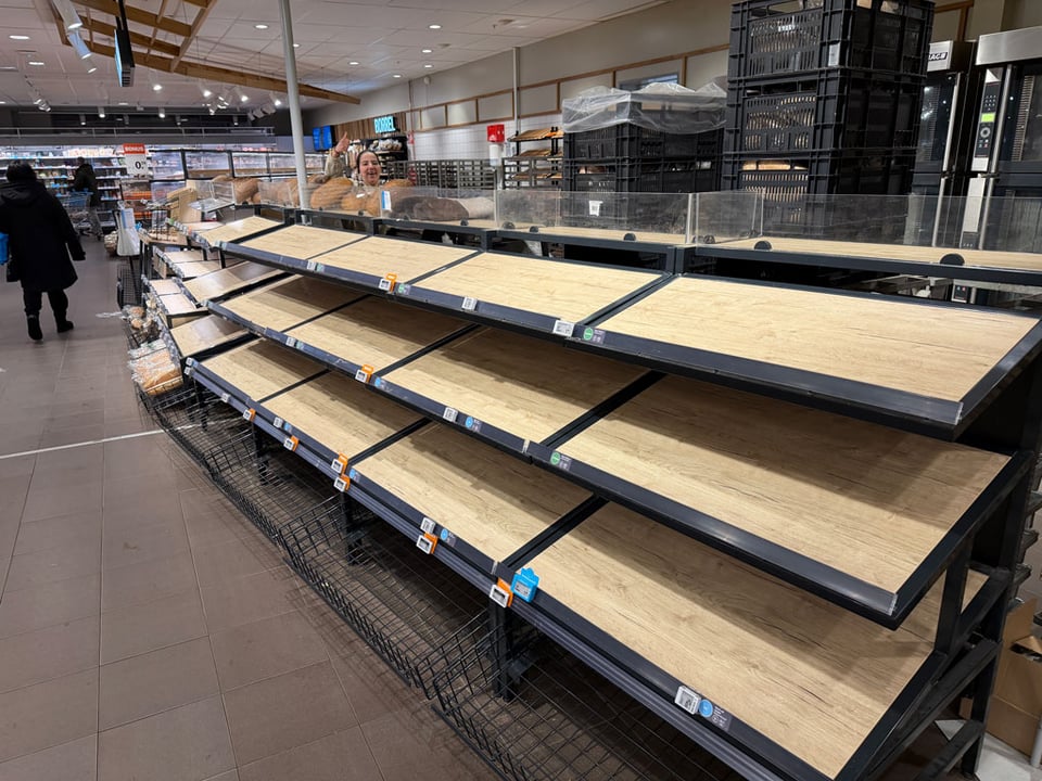 Empty breadshelves at Albert Heijn. The baker smiles and waves behind the counter in the distance.