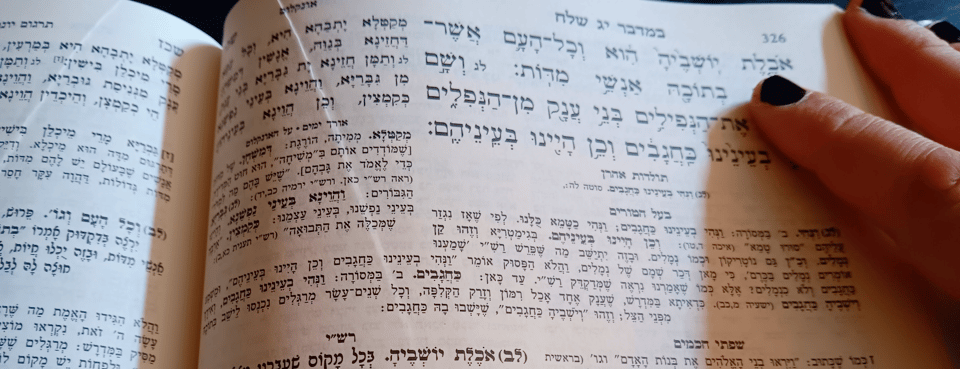 A picture of a page from a Torah with commentaries. A woman's hand is resting on the page, index finger (with black nail polish) pointing to the words that say "one who eats her inhabitants"