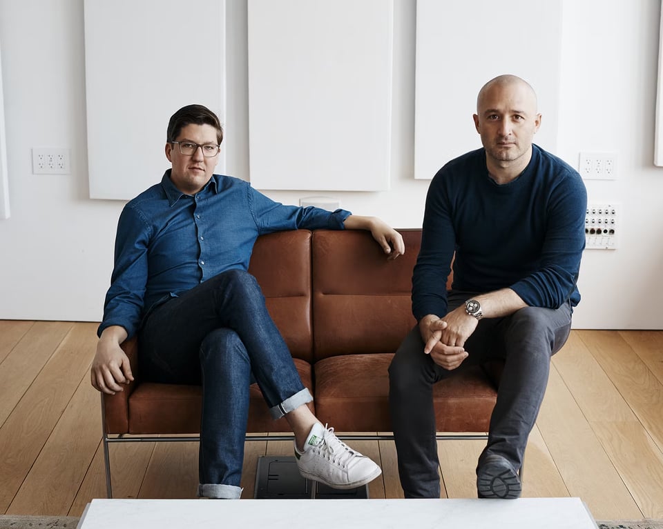 Two men sit on a brown leather couch in a minimalist white room with light wood floors. The man on the left wears glasses, a blue button-down shirt, and rolled-up jeans with white sneakers. The man on the right is bald, wearing a dark navy sweater and dark pants. Behind them are white walls with white panels and what appears to be a small control panel or device mounted on the wall.
