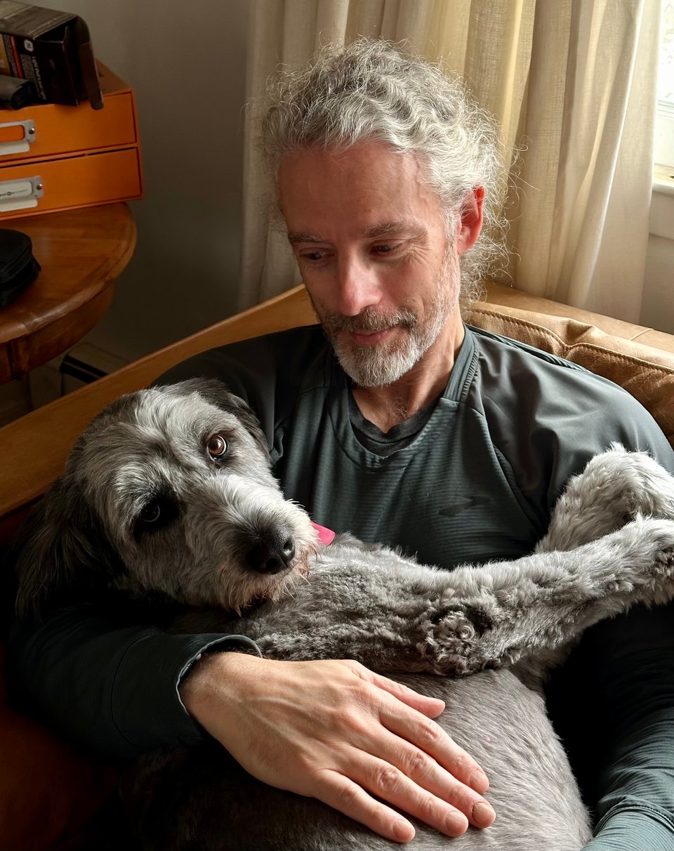 Photo of white man on a light-brown leather sofa, holding a reclining gray Aussiedoodle. He’s looking down at the dog, and she is looking at the camera with an exasperated “get me out of here” expression.