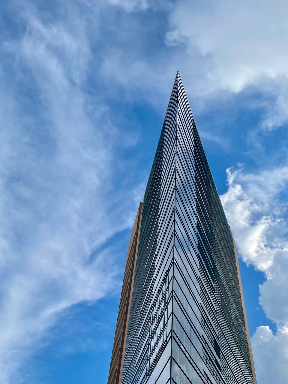 A glass building with a sharp edge exaggerated by the angle the photo was taken at. It sits in foreground of a bright blue sky with summer white clouds