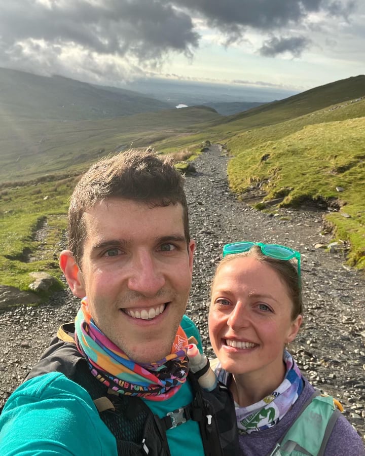Kelsey and Adam looking happy, standing along a gravel path in the mountains of North Wales. There are clouds in the sky, sun over their shoulders, and green grass to either side of the trail.