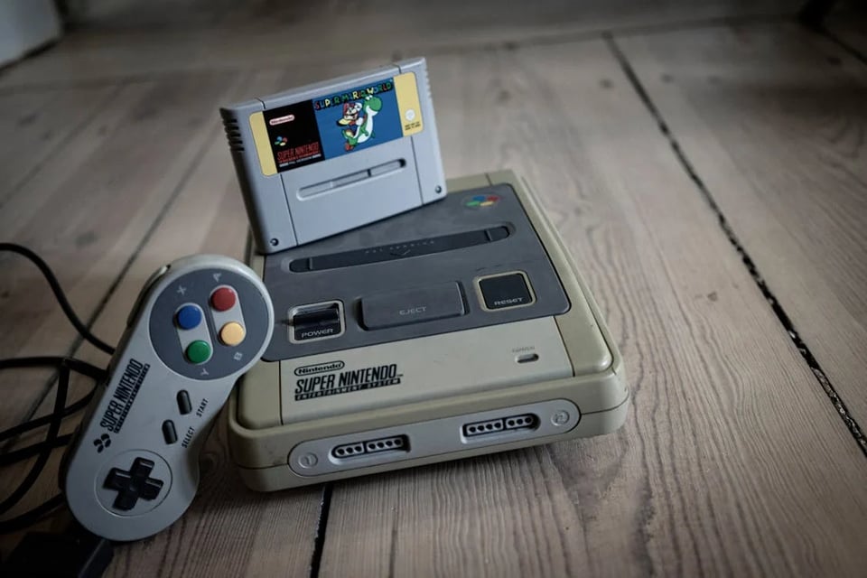 The photo of a super Nintendo system, with a cartridge perched on top of it and a controller, leaning against the side