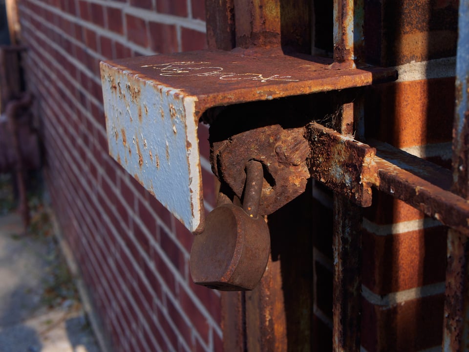 A rusty padlock, securing a gate. Red brick wall in the background.