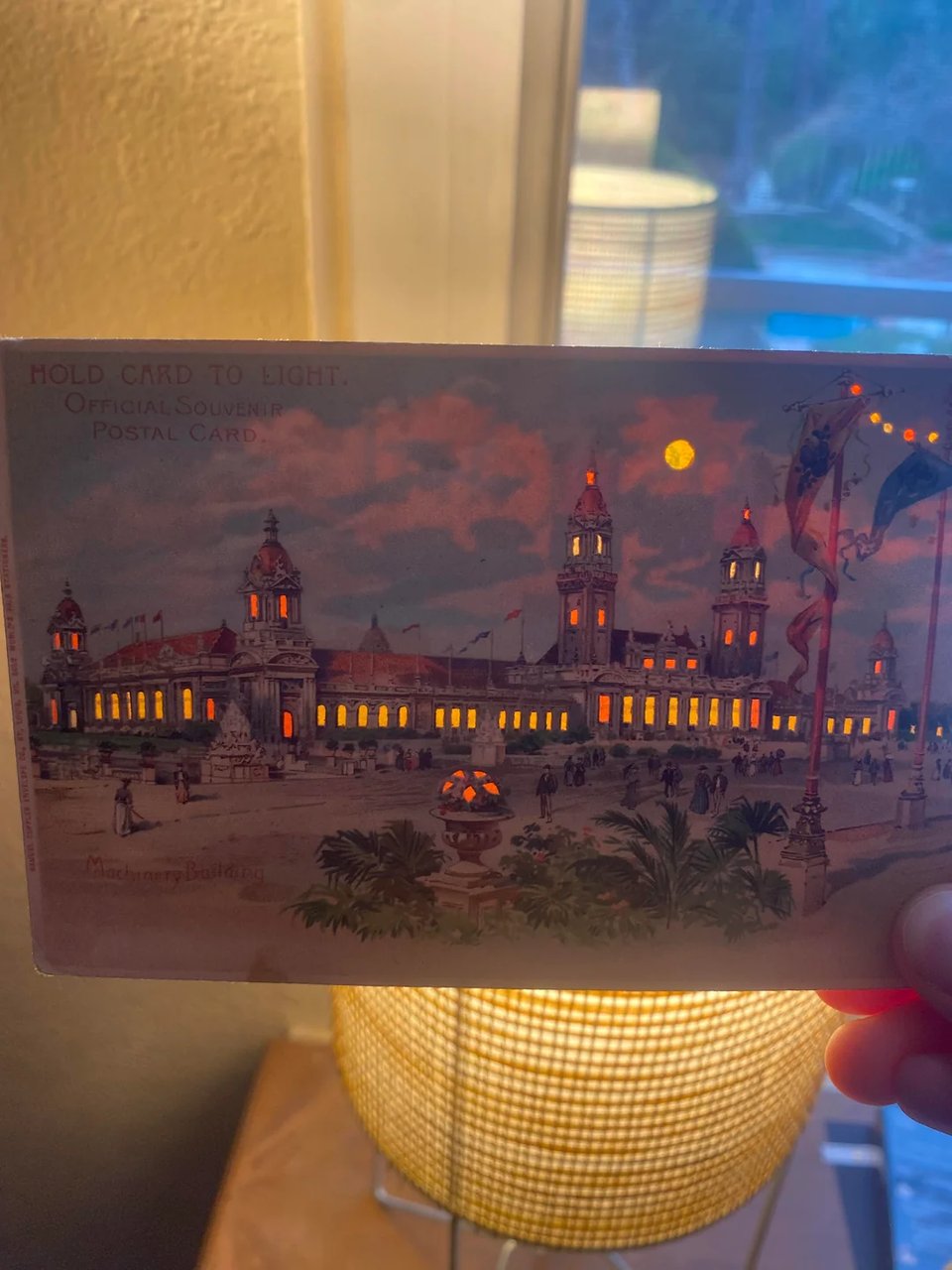 A hand holds up an old-fashioned postcard. It is a drawing of a large old 19th century grand building, and the windows are all glowing -- as if they were tiny colored holes in the postcard, through which light shines through