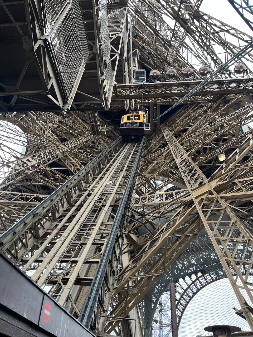 A shot of the Tour Eiffel from the inside, looking up. A web of metal with the yellow elevator and its rails in the middle.