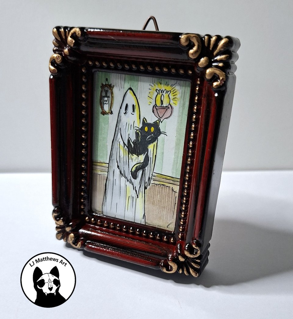 A small, intricate picture frame displays a whimsical illustration of a ghost holding a black cat in a room with a clock and a glowing light.