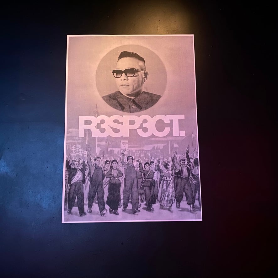 A poster shows a circular portrait of a man with glasses and dark hair at the top, with 'R3SP3CT' in large letters below. The bottom half features a black and white photograph of a crowd of people with raised fists in a solidarity gesture. The poster is photographed against a dark background with dramatic lighting.