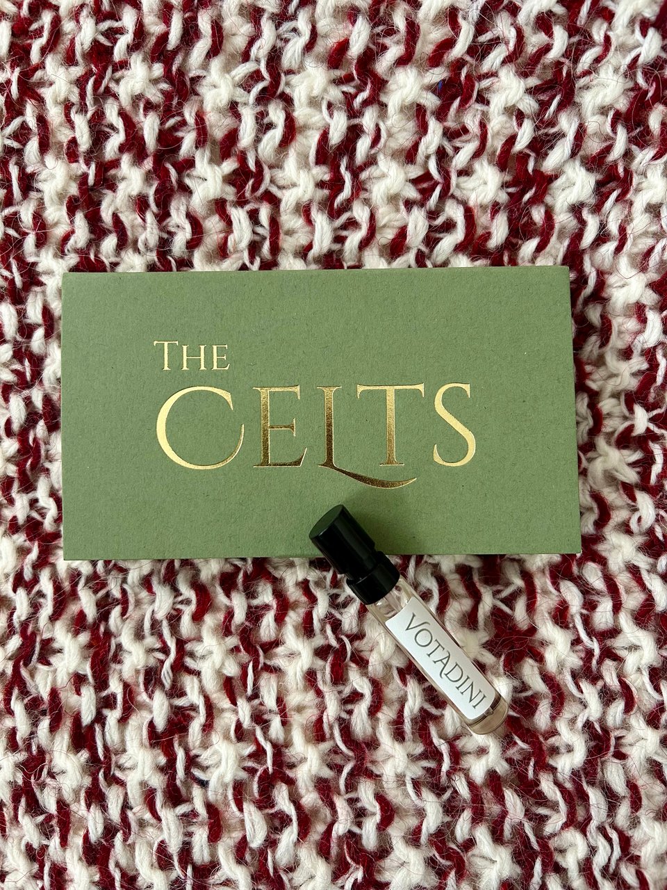 A small green box with golden writing 'The Celts' is on a red and cream knitted woolly cushion. There's a small glass vial of perfume resting on the box, called Votadini. Image by Rowan Ambrose.