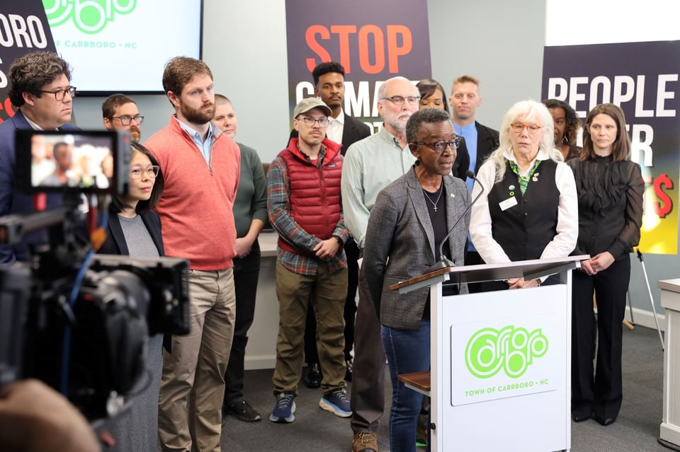 A short black woman with short cropped hair and a gray blazer stands in front of podium, surrounded by other people and signs about climate change as well as a video camera recording her in close crop
