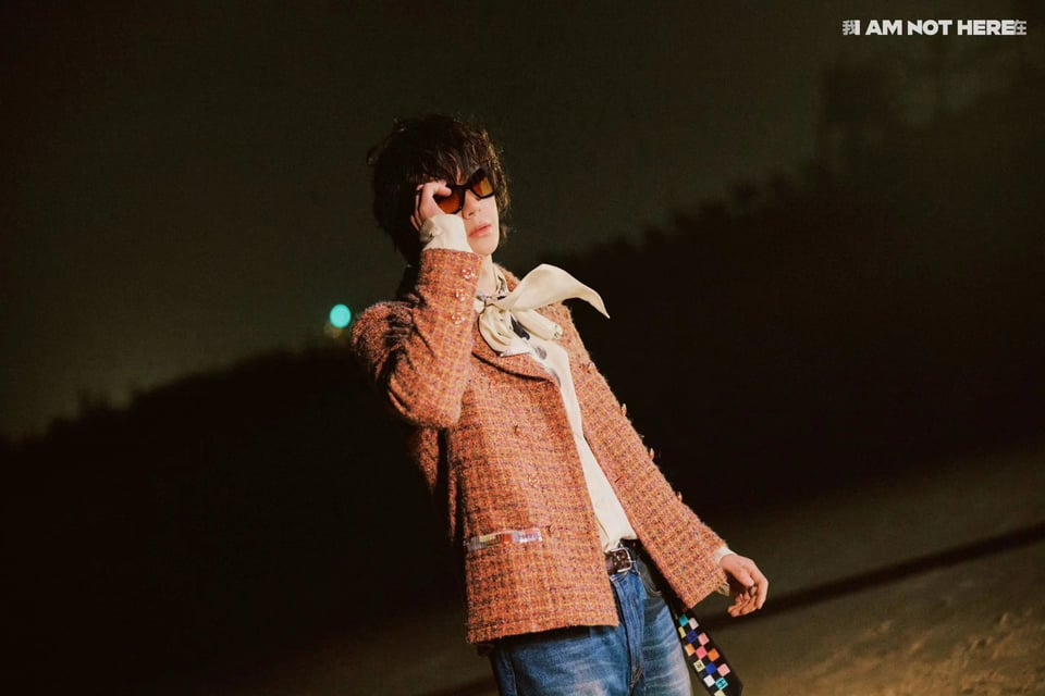 Wang Yibo in the incredible suit he wore for his 2024 New Year's Eve performance. A light orange-red Chanel jacket over an off-white shirt (probably silk) with a giant pussy bow, a pair of blue jeans with a colorful scarf tied as a belt, a shag wig, and sunglasses. At night. The whole look is faintly Bob Dylanesque.