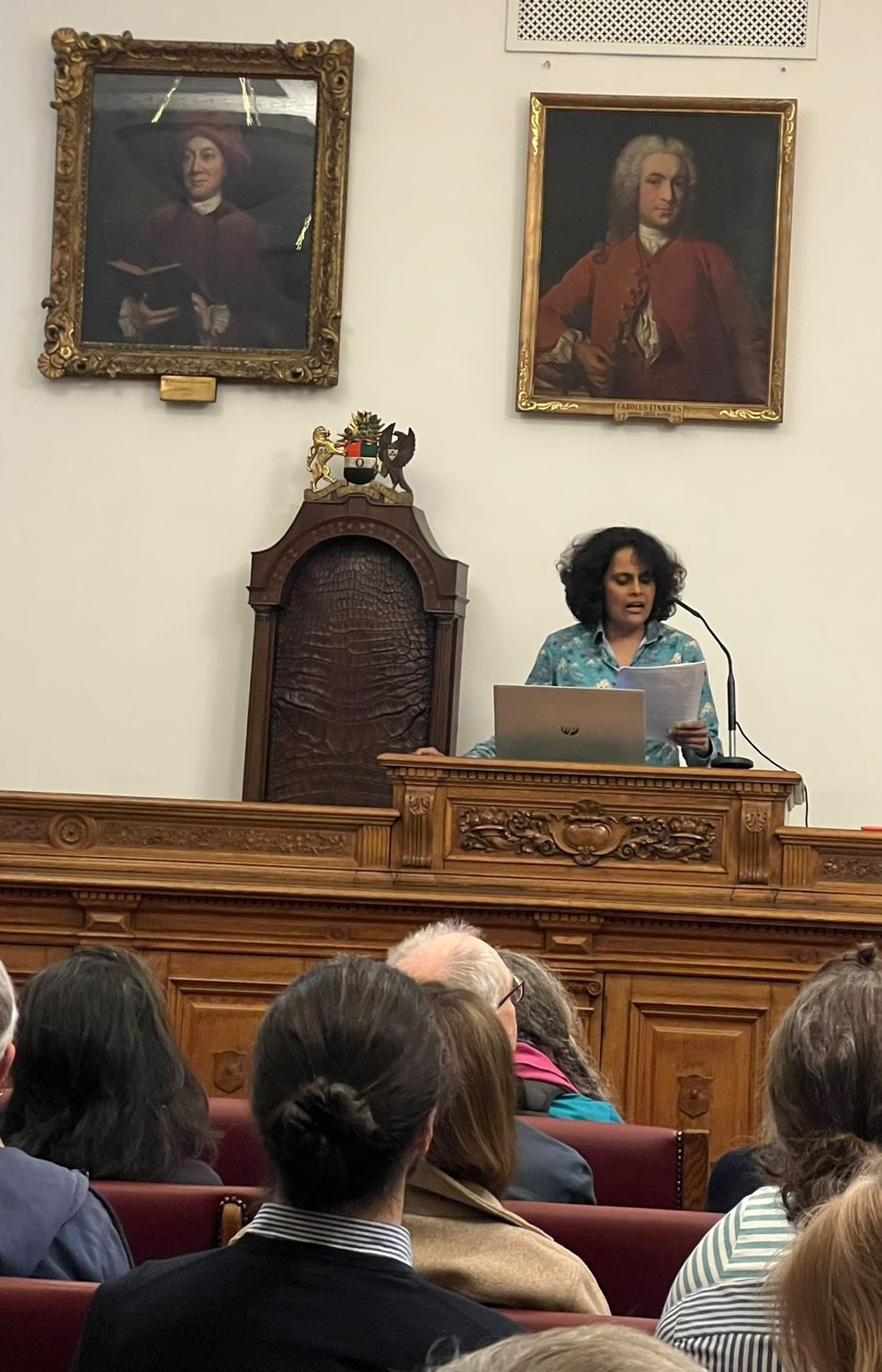 Me in a blue and patterned collared shirt,  speaking at a wooden lecture podium, laptop in front of me and lecture printout in hand. Behind me are two historic portraits (Carl Linnaeus directly above me). To my right is a tall, narrow, wooden chair (its leather back is visible) with a coat of arms on top. In the foreground are the heads of the packed audience, who appear to be listening intently.