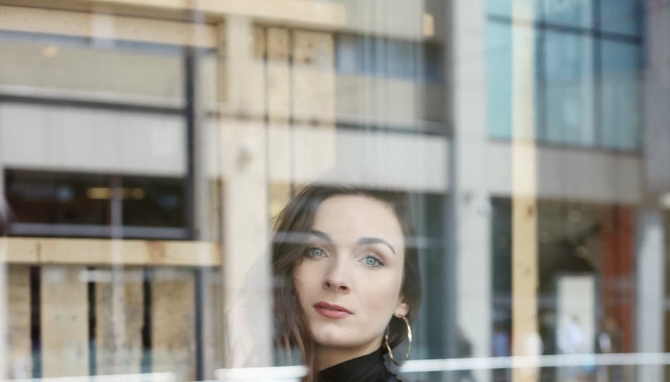 A portrait of Claire reflected in a window with a background of modern buildings.