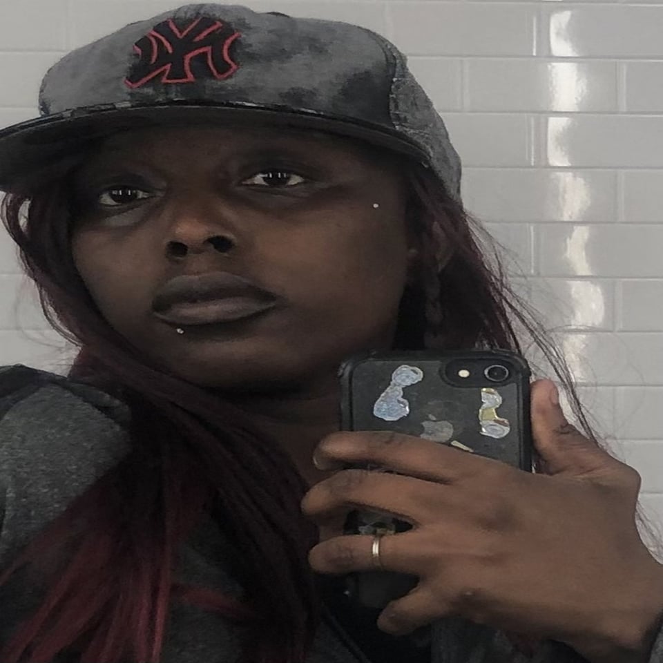 a person with dark red hair and a black baseball cap takes a selfie in a white tiled room