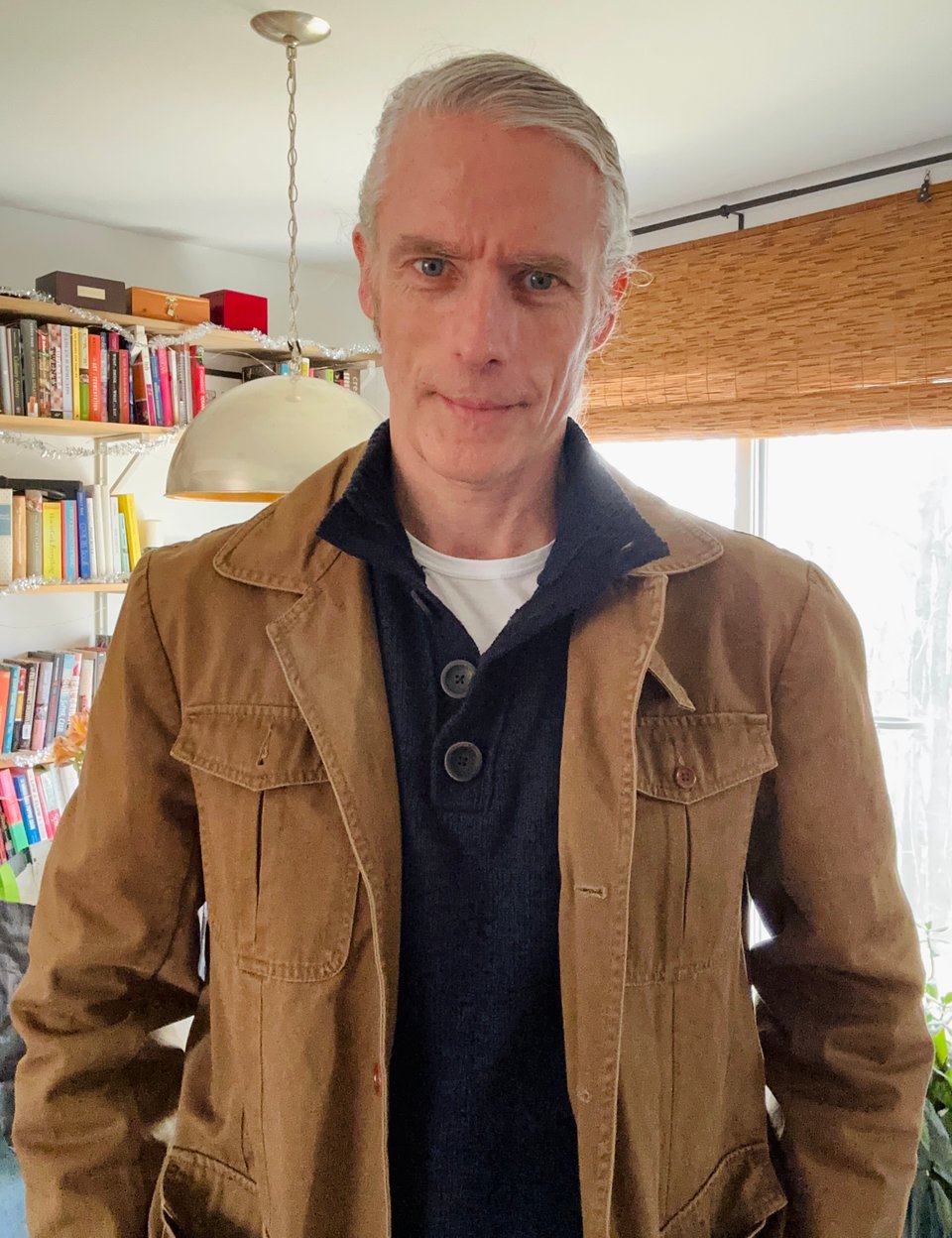 photo of white man in light brown outdoor coat over navy sweater with a button-up standing collar and white T-shirt. His hair is slicked and tied back so he looks somewhat professional. There are shelves of cookbooks behind him, and 3 wooden boxes on the top shelf, containing the ashes of his 3 greyhounds.