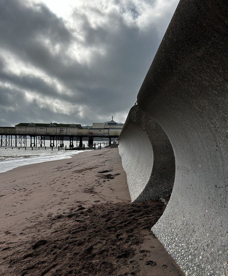 A huge sea wall curving over a sandy red beach. In the distance is a Victorian pier. There's a gap in the wall and lots of footsteps and pawprints of people coming on and off the beach. At the base of the curve, the sand mix in the concrete has been steadily scoured away, revealing the aggregate mix. This reflects the light more than the concrete that hasn't been worn.