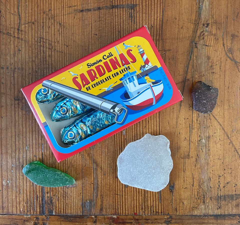 A box of blue foil wrapped chocolate sardines is on a battered antique pine trunk. There are pieces of green brown and clear seaglass around the box. Image by Rowan Ambrose.