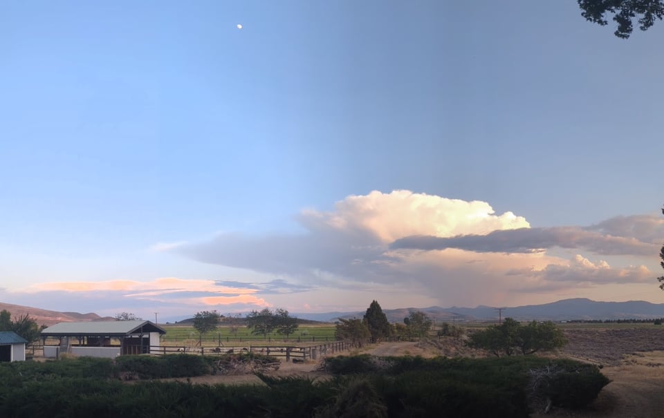 A panorama of a thunderhead waning over a mountain range, with a smudge of moon up top.
