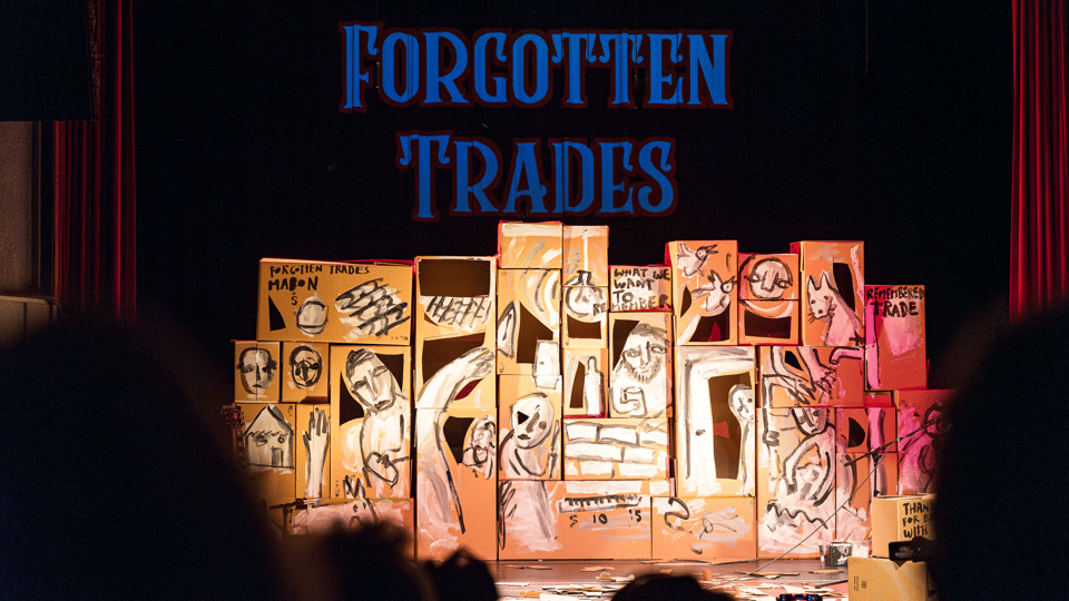 Cardboard boxes painted on with multiple figures, and the title 'Forgotten Trades' displayed in a live theatre show.