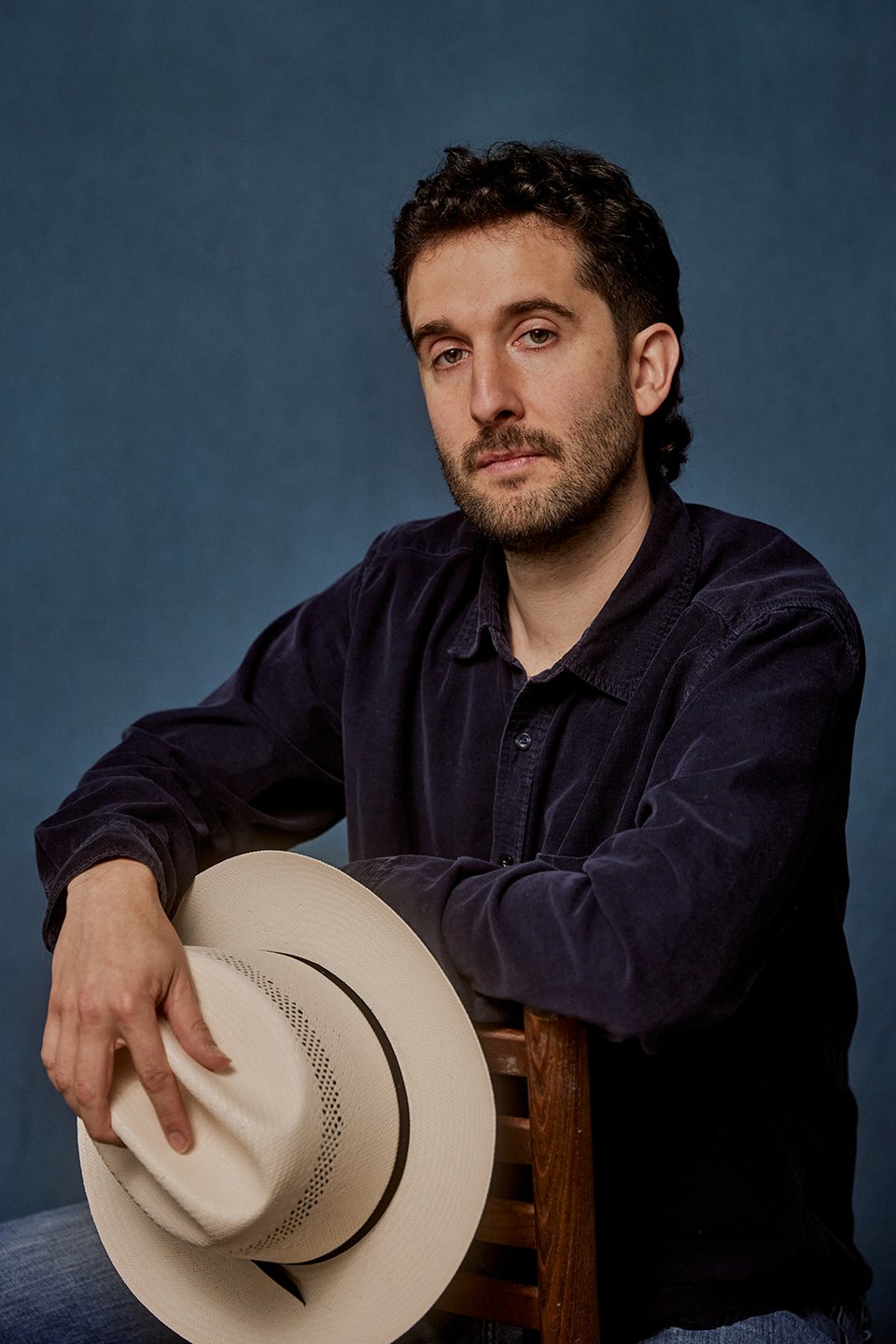 A man with a light beard sits straddling a chair and holding a cowboy hat in his right hand.