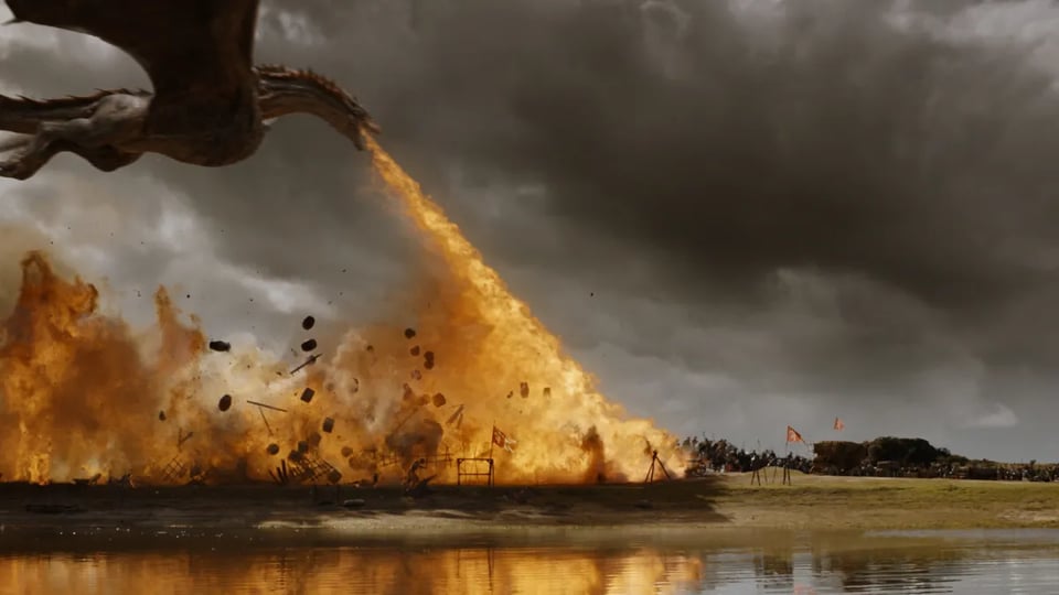 A still image from Game of Thrones, the TV show, showing a dragon flying above land and strafing and burning teh land with a long flame from its mouth. A few small flags are visible, suggesting that the dragon is attacking a military formation