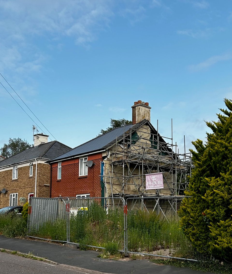 A blocky semi-detached house which has become detached because the other half has been demolished. The party wall has waterproof membrane and scaffolding. The plot is fences off and thick with weeds.