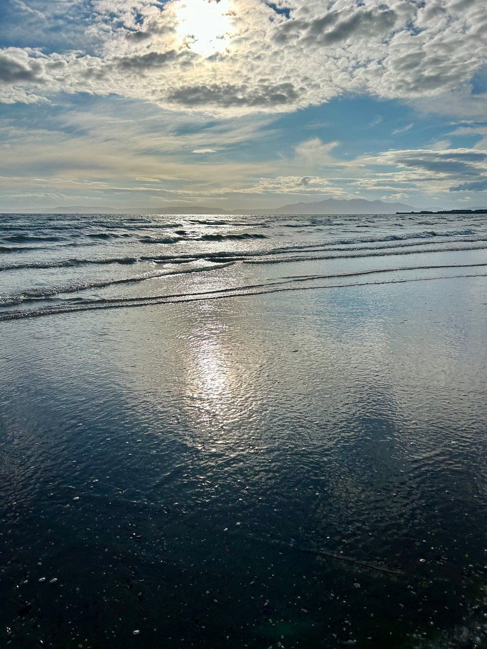 A beach scene in Scotland. There are distant mountain peaks, part hidden in mist, puffy white clouds in a blue sky, and waves gently rolling in. Image by Rowan Ambrose.