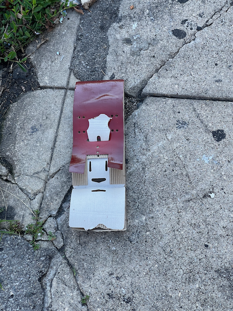 A cardboard backing board for some retail product is torn open, lying on a cracked sidewalk. The hole for the retail hanger is placed so it looks like a friendly mouth, two slots above it look like eyes, and two strips of cardboard on either side of that face look like hair. Red paper overlaying that cardboard makes it look like a hat.