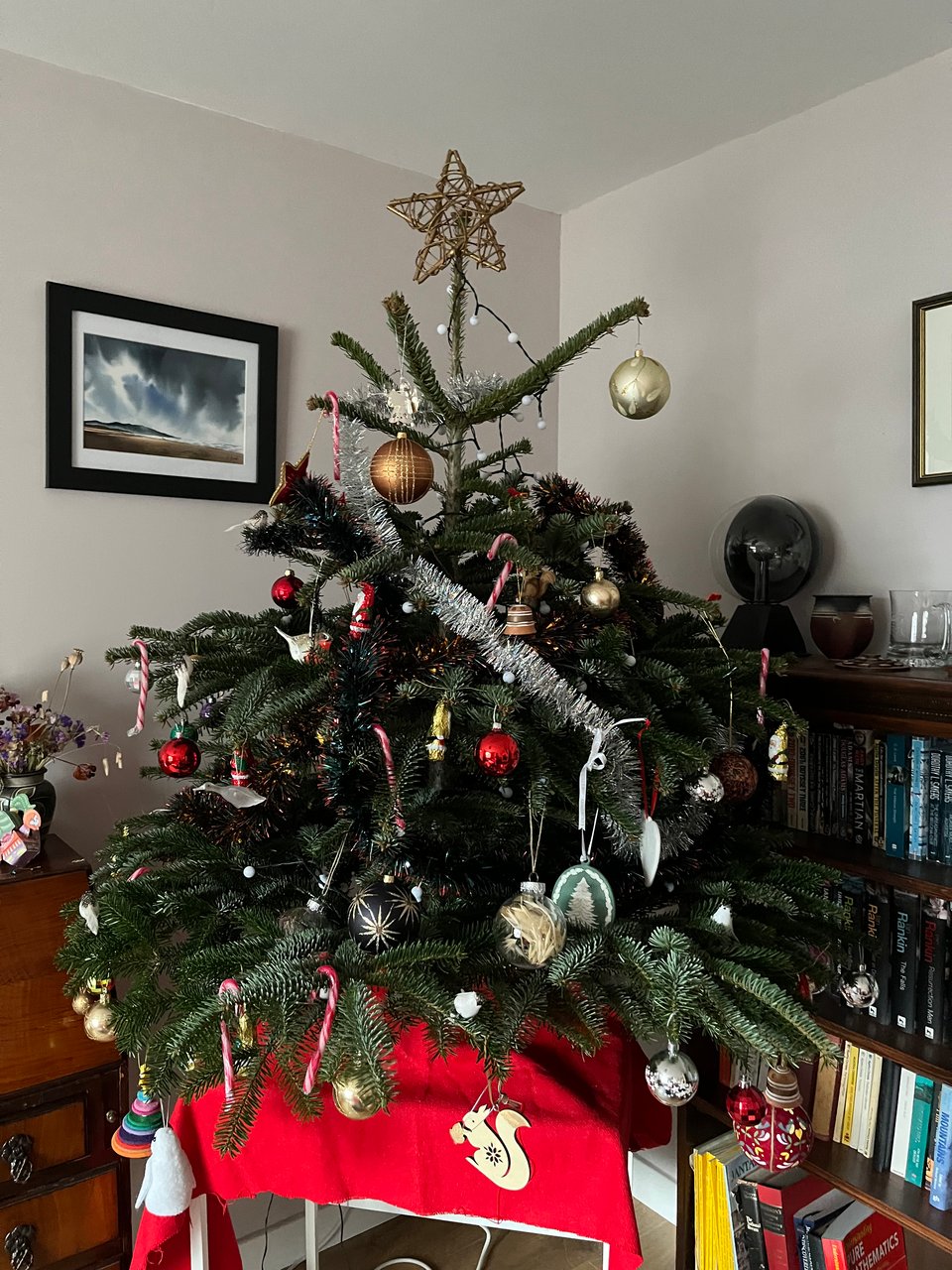 a decorated christmas tree on a red cloth. It is decorated with baubles, candy canes and tinsel. There is a squirrel ornament dangling in the foreground and a goose flying to the left