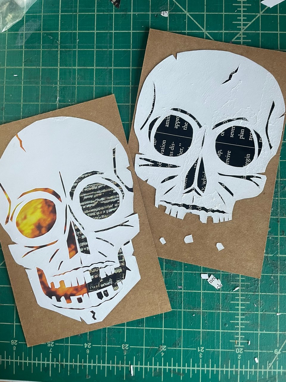 Two postcards featuring papercut skulls sitting on a green cutting mat.