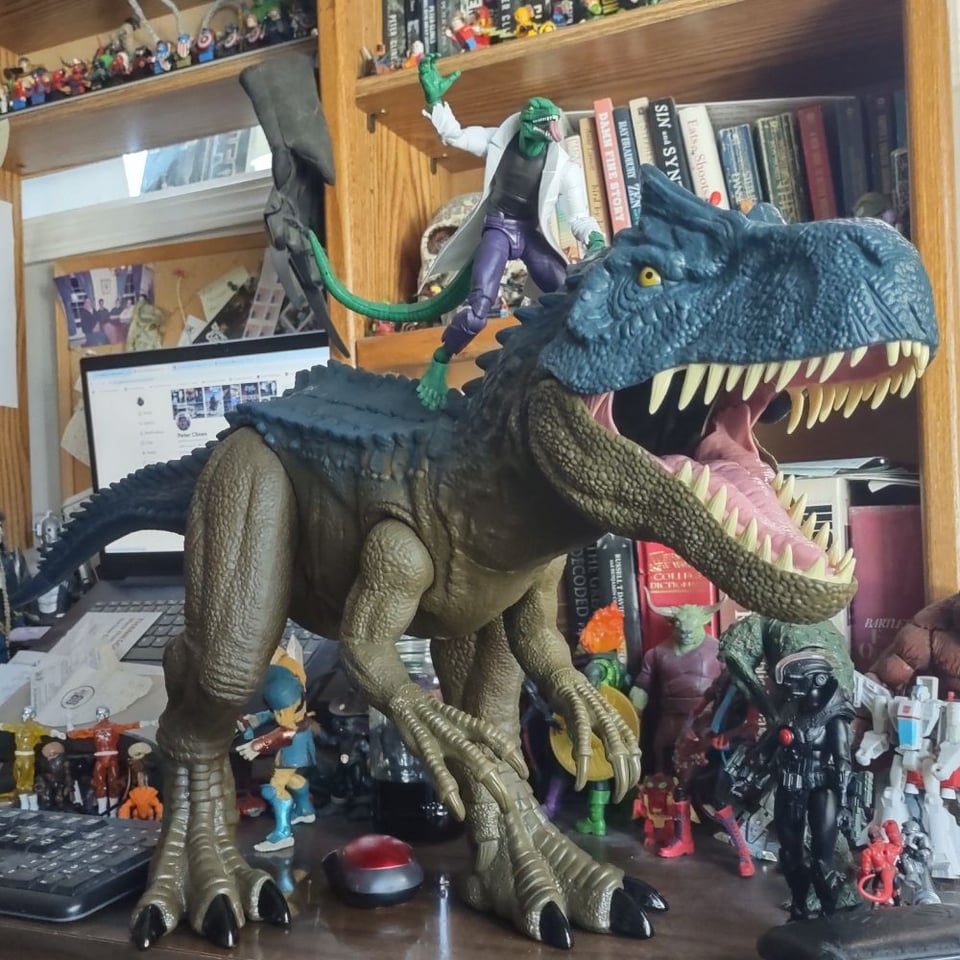 A huge Allosaurus toy stands on a wooden desk, dwarfing all the action figures around it (and the one riding it). A laptop computer can be seen in the background, showing a Bluesky page.