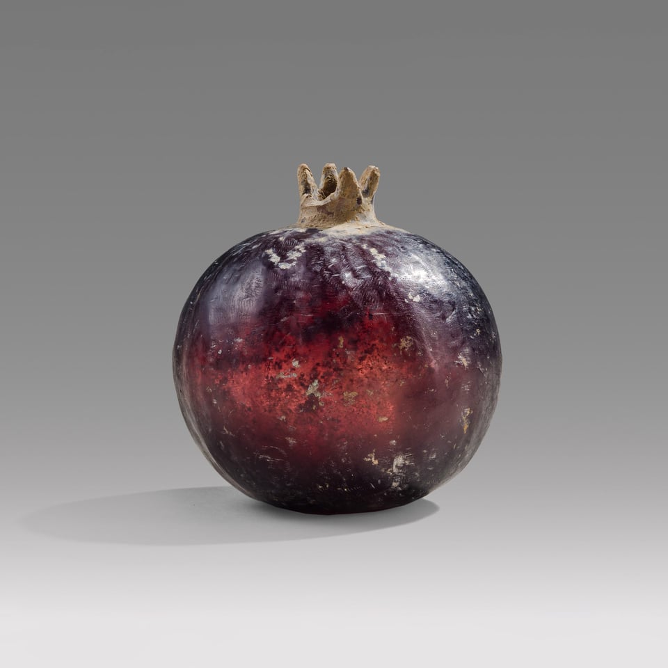 On a grey background, a perfect purple glass sphere glows with red and violet and deep purple hues. A tiny crown in earth colors hints that this is a pomegranate. Small scuffs and dirt veil the surface, but in no way mar the smooth lines or naturalistic beauty of the piece.