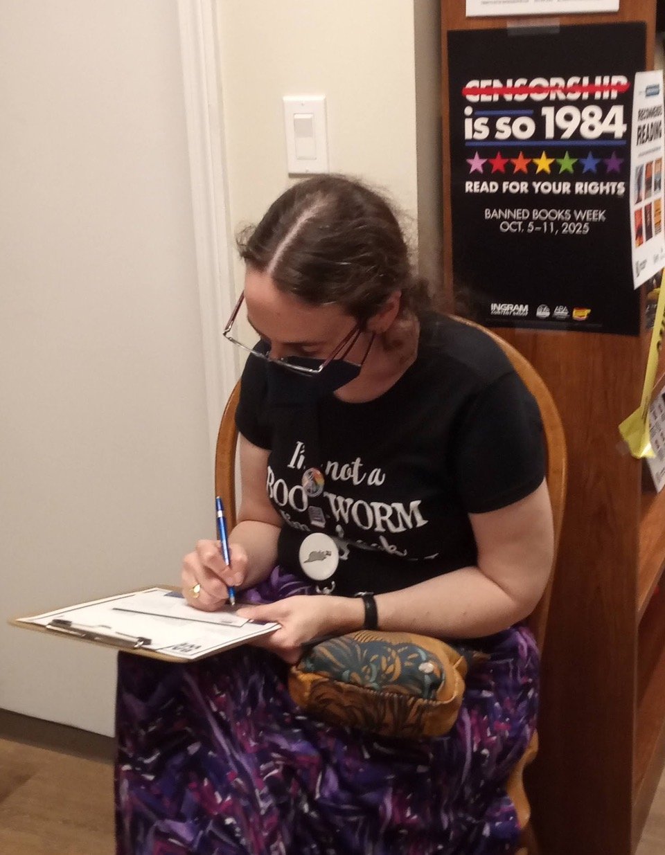 The author sits in a chair, writing a postcard on a clipboard balanced on her knee. There is an anti-censorship poster in the background.