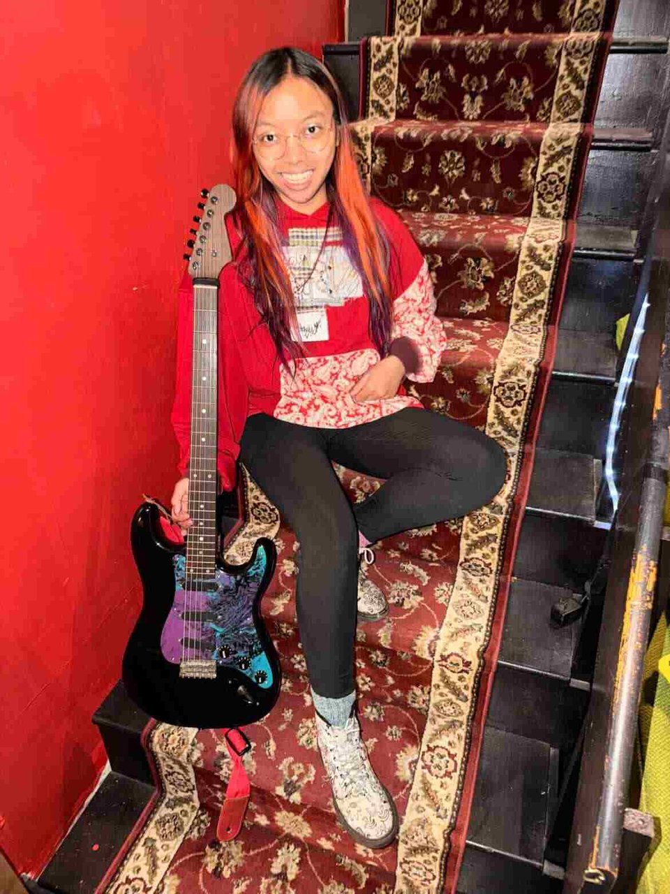 A woman with long hair dyed red wearing a red top and black leggings and holding a black Strat-style guitar with a colorful blue, purple and black pickguard sits on carpeted steps against a red wall.