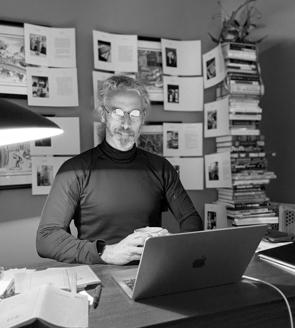 b/w photo of white man at writing desk, smiling a little. He's holding a cup of coffee with a laptop in front of him. He's wearing round glasses and the light is reflecting off them so you can't really see his yes. In the background there are sheets of paper taped to the walls; they have photos and text on them