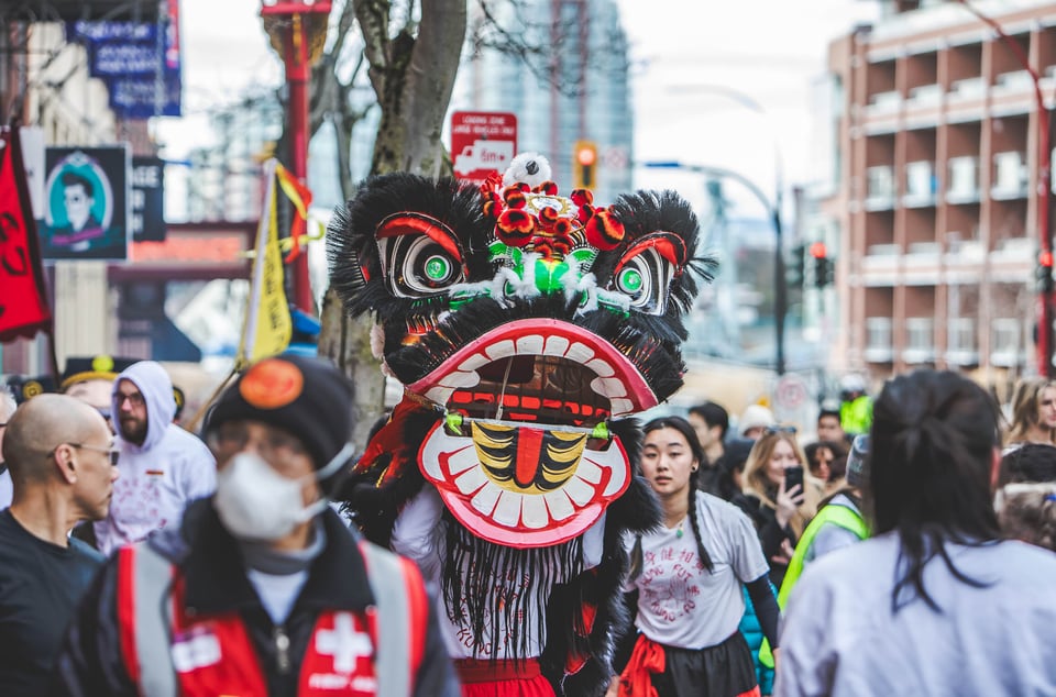 A lion head with black trim and green eyes being carried by a person in a Hung Fut Kung Fu uniform, in a crowd in Old Town Victoria