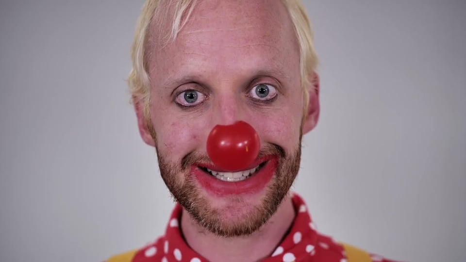 Simon stares with dead eyes into the camera, wearing a red nose, clown lipstick and an insincere smile