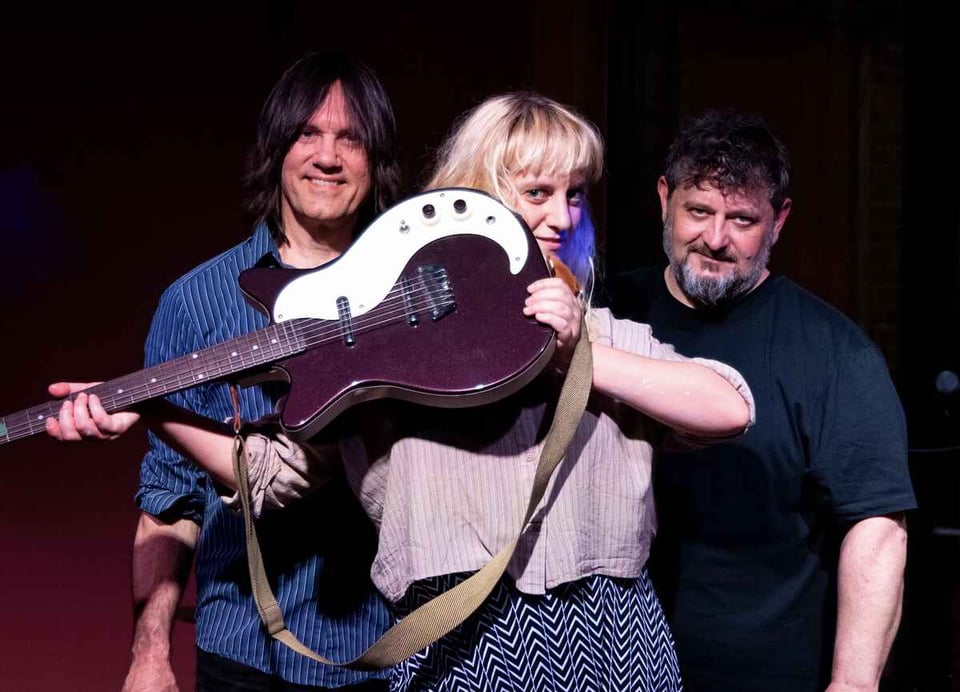 A woman with blond hair peers around the body of a guitar she's holding up in front of her. She's flanked by two men, one clean-shaven and wearing a blue striped button-up shirt and the other with a beard wearing a dark-colored t-shirt.