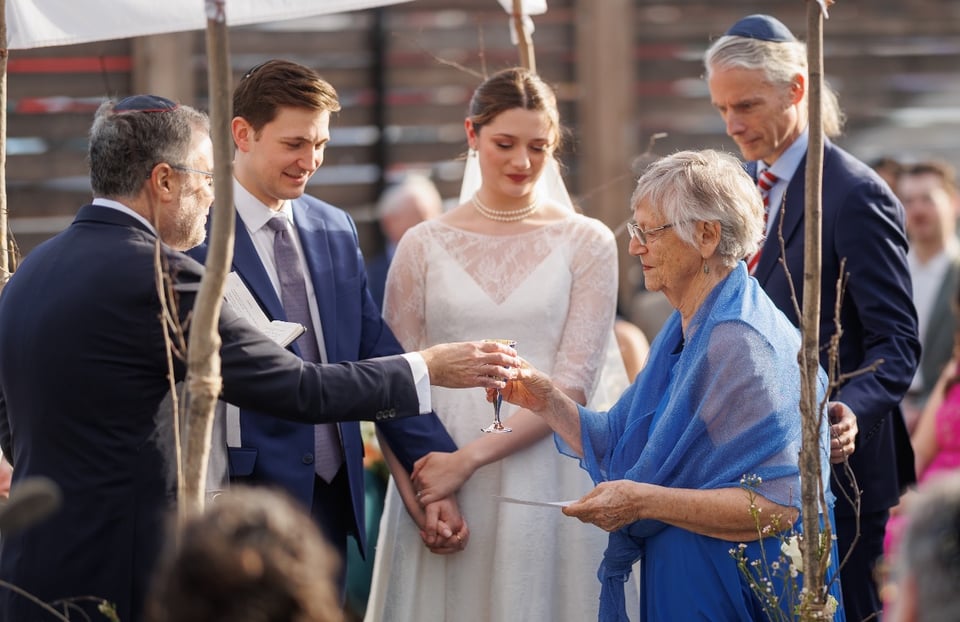 Photo of Jewish wedding, showing grandmother in blue dress handing a wine-glass to the rabbi under the chupa, while her son, the bride, and the groom look on. The bride is holding the groom’s hand and she looks beautiful.