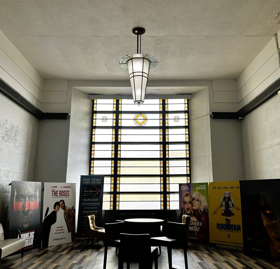 A double height lobby with a stunning stained glass window that playfully hints at being strips of film. There's a great Deco ceiling light, and artex-patterned walls. There are large card posters for films. There are no people.