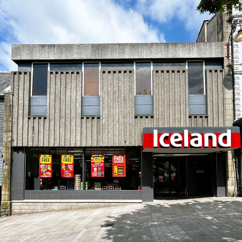 A brutalist supermarket front on a sloping pedestrianised street. The ground floor is mostly large windows with dark square tiles. Above the entrance is a heavy cantilevered porch roof. Above street level the building is clad in concrete that has a vertical rib pattern. Four long window recesses are symmetrically set into this. The inner structure rises above the concrete cladding, and is then capped with a smooth concrete parapet.