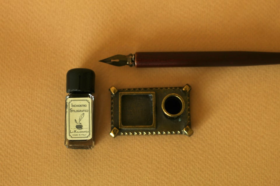 A nibbed pen, a bottle of ink, and an inkwell all sit against a yellow fabric background.